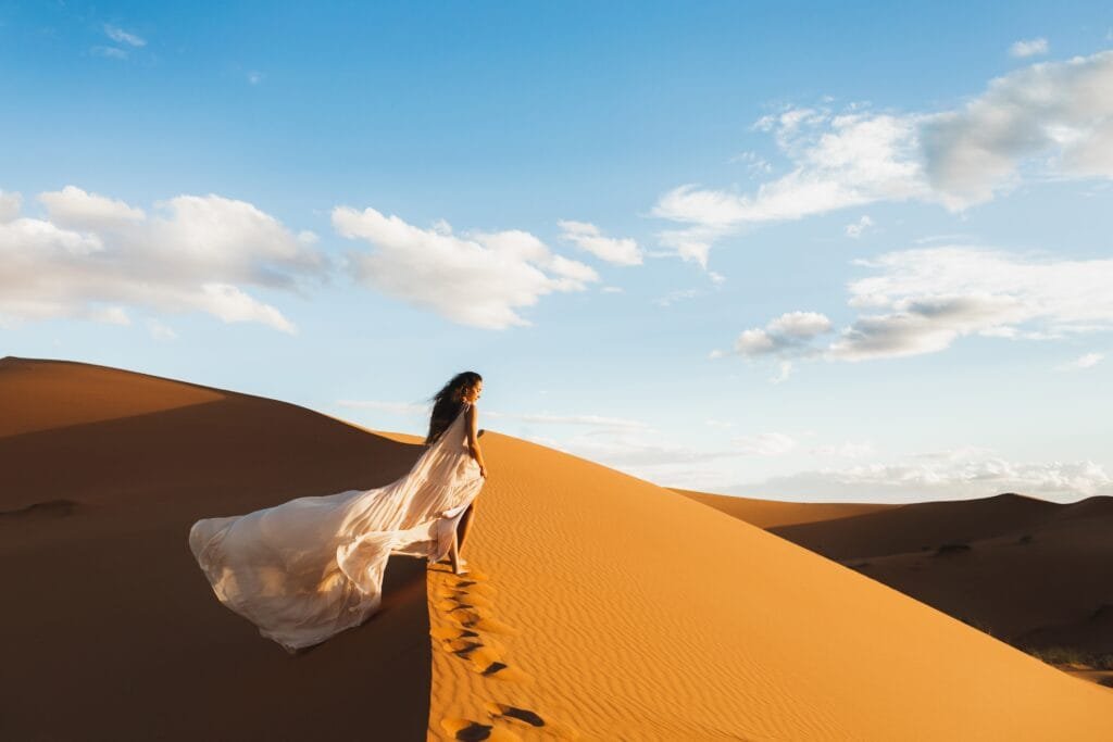 Woman in amazing silk wedding dress with fantastic view of Sahara desert sand dunes in sunset light. Landscape of Morocco, Africa.