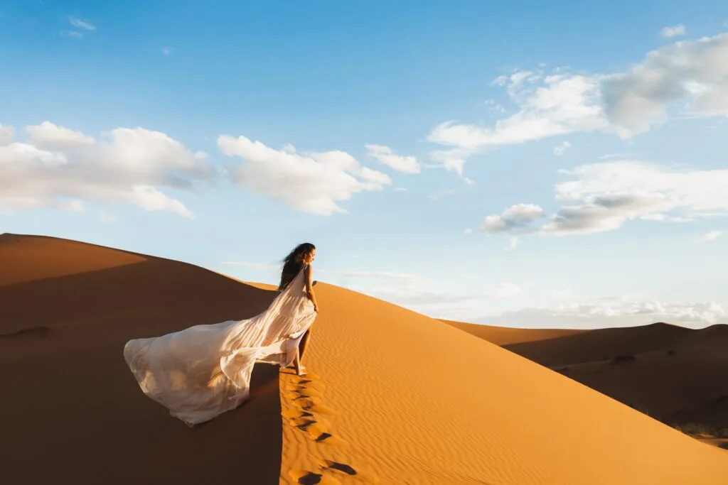 Woman in amazing silk wedding dress with fantastic view of Sahara desert sand dunes in sunset light. Landscape of Morocco, Africa.