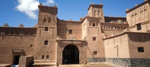 Majestic earthen towers of Kasbah Amridil rising above the lush palm oasis of Skoura in southern Morocco, with traditional pisé mud-brick architecture glowing in warm sunlight against a clear blue sky and the distant Atlas Mountains on the horizon.