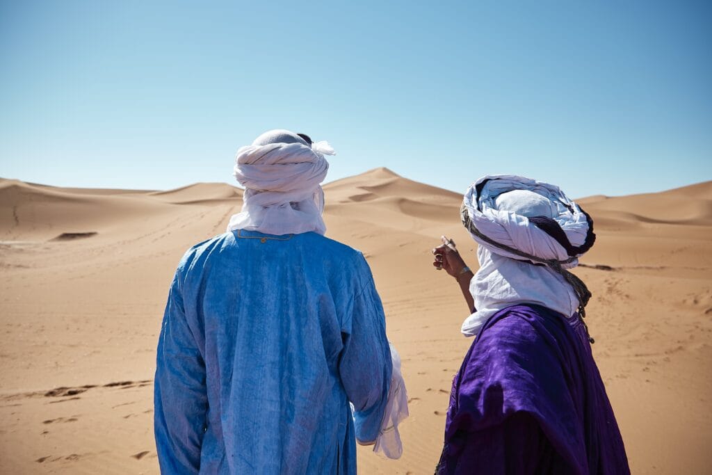 A horizontal shot of two friends standing in front of Chigaga dunes in the Moroccan desert on a sunny day