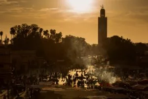 Vibrant Djemaa el-Fna square in Marrakech, Morocco, bathed in warm golden sunset light, with smoke rising from open-air food stalls, crowds of locals and travellers filling the plaza, and the iconic Koutoubia Mosque minaret rising above the rooftops in the background.