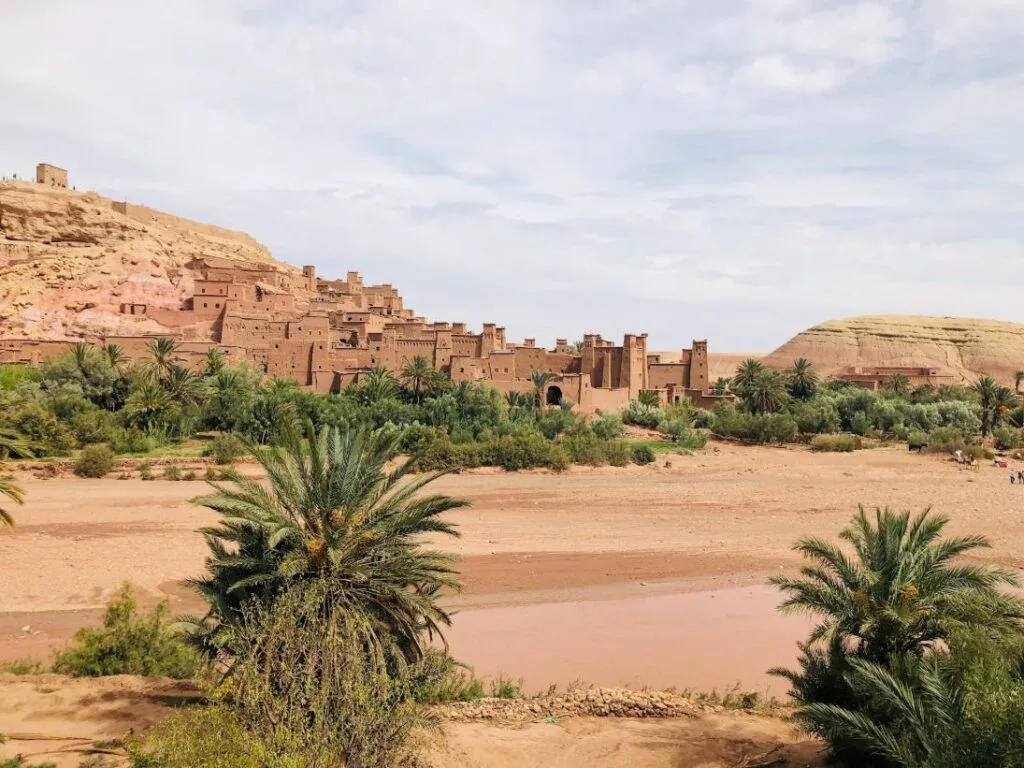 The ancient UNESCO-listed ksar of Aït Benhaddou in southern Morocco, its tiered earthen kasbahs and defensive towers built from sun-dried red clay and straw stacking dramatically up a rocky hillside above the dry Ounila riverbed, set against a vast blue sky near the city of Ouarzazate on the edge of the Sahara.
