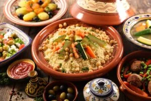 A spread of traditional Moroccan dishes featuring a large plate of fluffy steamed couscous topped with vegetables and tender meat, alongside a conical clay tagine pot filled with slow-cooked lamb or chicken with olives, preserved lemons, and aromatic spices, garnished with fresh herbs and served on a decoratively patterned table.